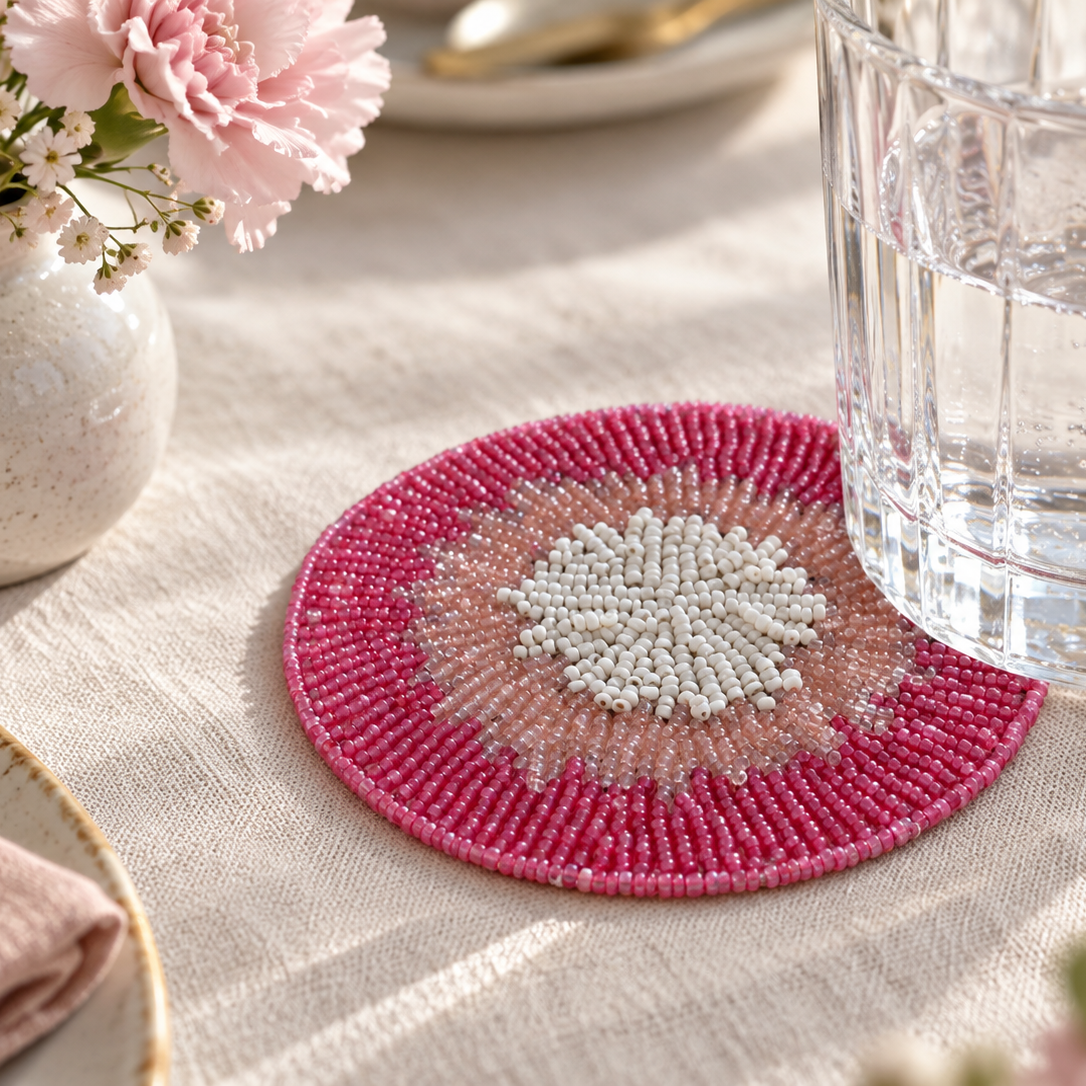 A round beaded placemat with a pink and white geometric pattern, styled on a beige linen tablecloth next to a glass of water and a small vase of flowers.