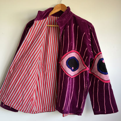 Purple velvet jacket with eye designs on a hanger against a white background