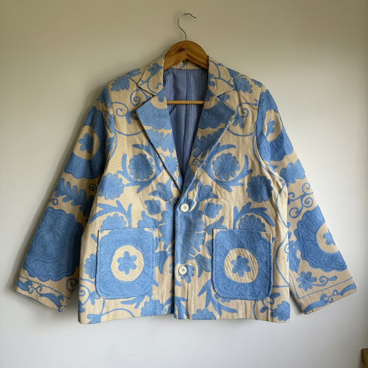 Blue suzani embroidery Jjacket on a hanger against a plain wall.