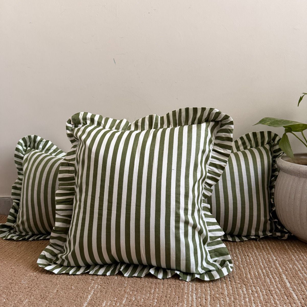 striped throw pillows with ruffled edges in olive green and white, arranged on a woven beige rug against a plain wall with a potted plant partially visible on the right.