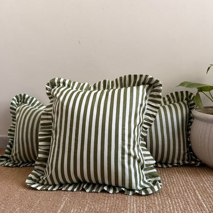 striped throw pillows with ruffled edges in olive green and white, arranged on a woven beige rug against a plain wall with a potted plant partially visible on the right.