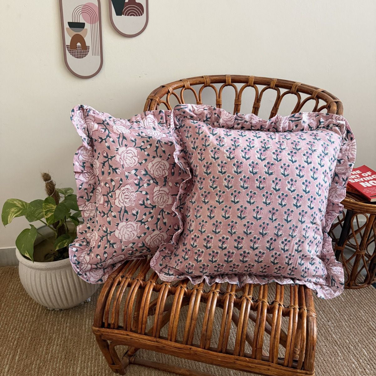 Two pink floral throw pillows with ruffled edges displayed on a brown wicker chair in a styled room setting.